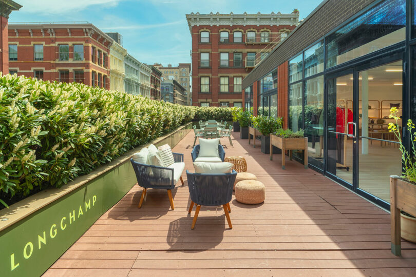 Outdoor wooden deck with lounge chairs, potted plants, and tables beside a building with large glass windows; city buildings visible in the background.