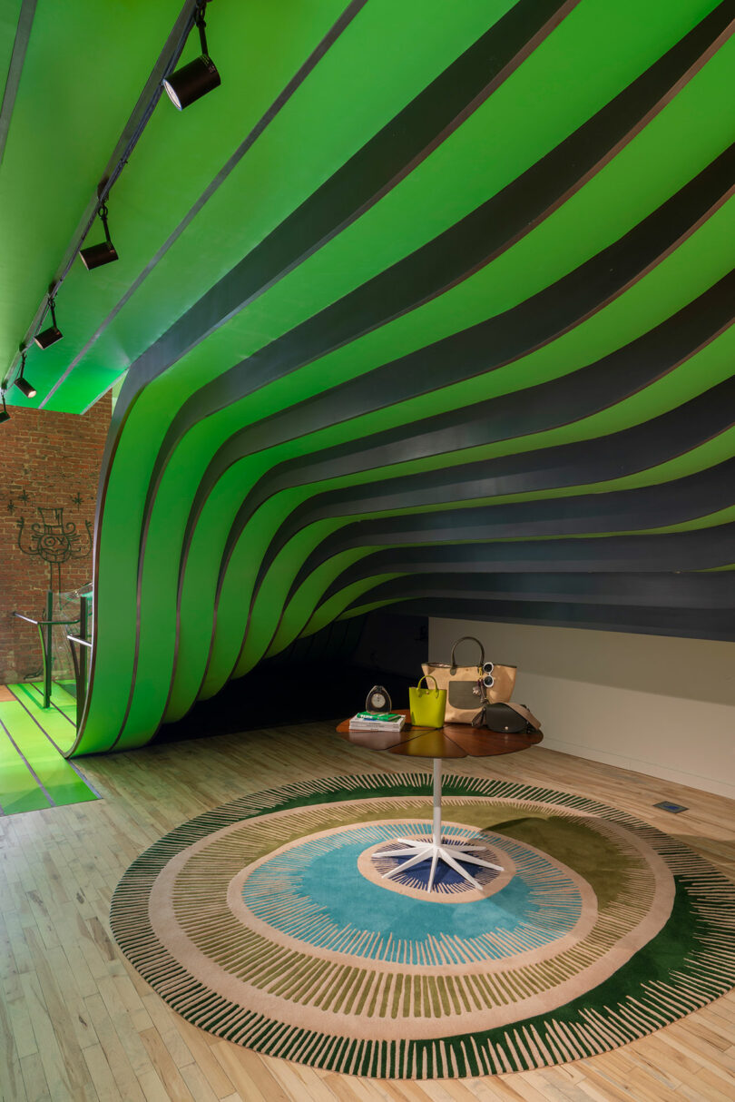 A modern interior with wavy green ceiling panels, a round patterned rug, a small round table displaying handbags, and wooden flooring.