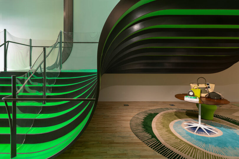 Modern interior with a curved staircase featuring green accent lighting, a round table with bags and books, and a patterned rug on wooden flooring.