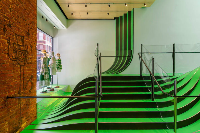 Modern store interior with wavy green and black staircase, exposed brick wall on the left, mannequins in front of large window, and glass railings along the stairs.