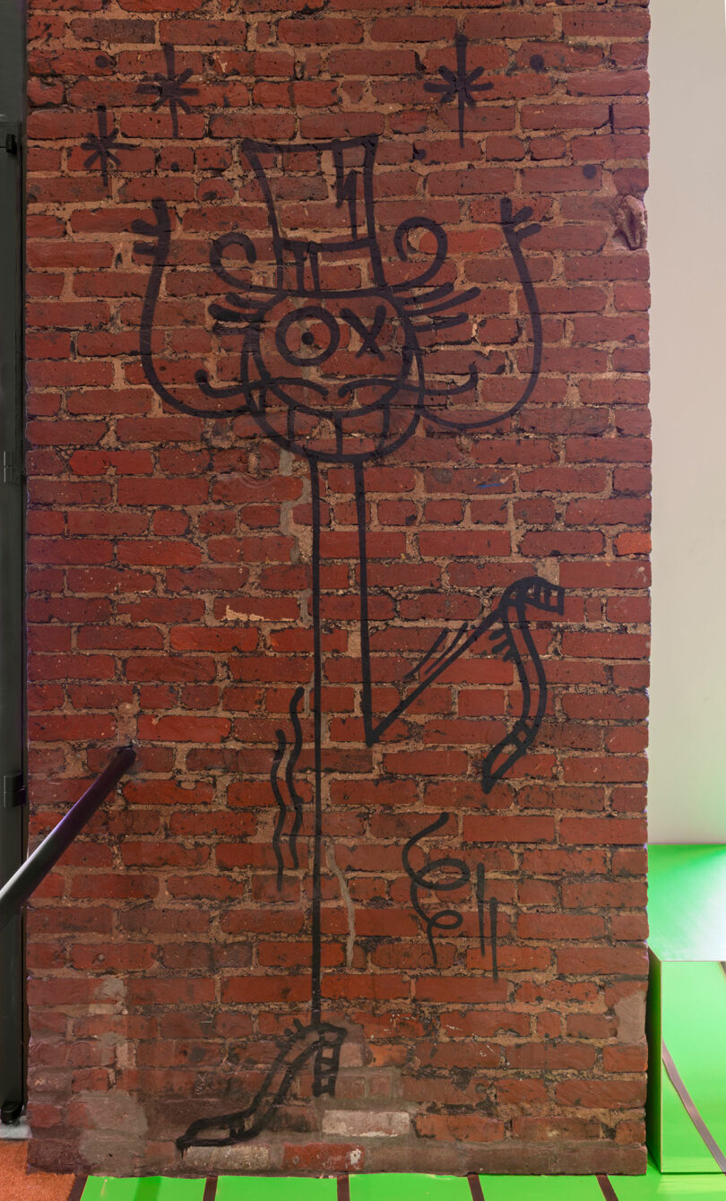 Black marker drawing of a whimsical, one-eyed figure wearing a top hat and high-heeled shoe on a red brick wall, with stars above and wavy lines beside it.