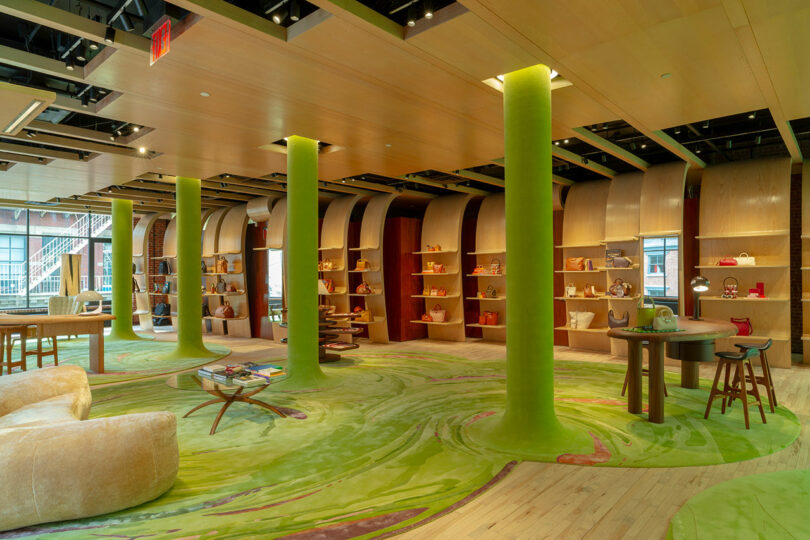 Modern retail store interior with green columns, wooden shelves displaying various handbags, and curved furniture on a green patterned floor.