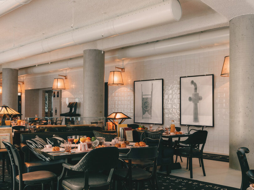 A modern restaurant interior with set tables, black chairs, concrete columns, framed abstract art on white tiled walls, and warm ambient lighting.