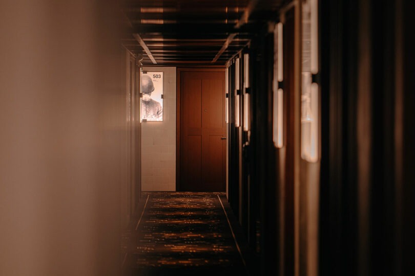 Dimly lit hallway with closed doors on both sides, soft warm lighting, and a sign marked "503" next to a blurred figure behind frosted glass.