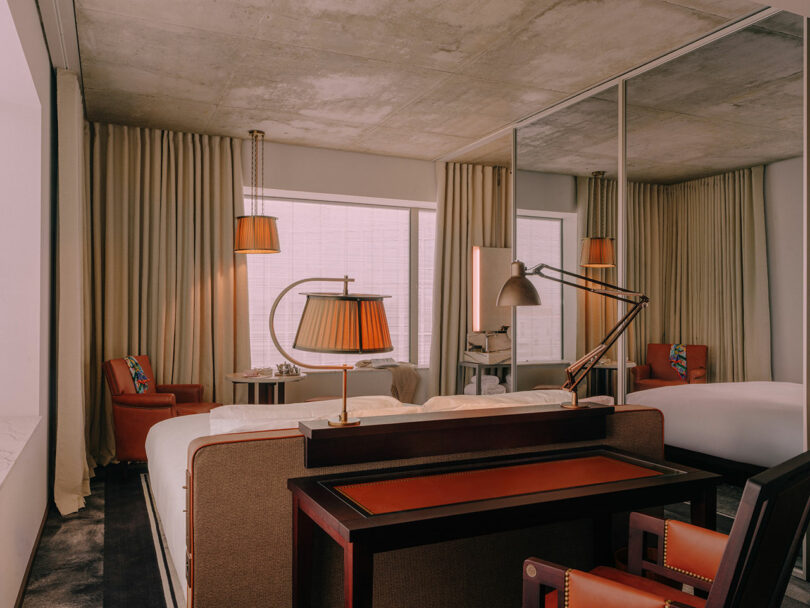 Modern hotel room with two beds, a work desk, two armchairs, large windows with curtains, mirrored wall, and industrial-style ceiling.