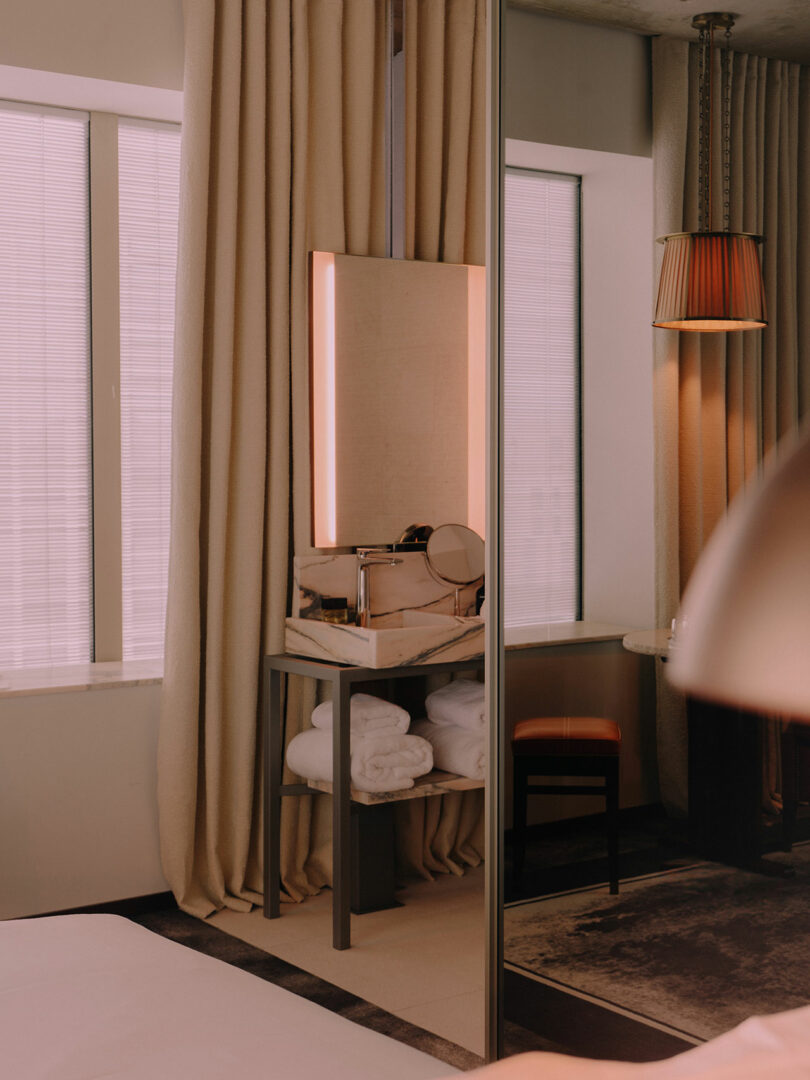 A vanity table with a backlit mirror, folded white towels, and a round mirror sits by curtained windows next to a cushioned chair in a softly lit room.