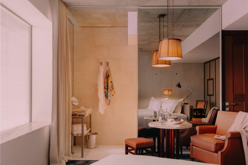 Modern hotel room with a made bed, leather chairs, a small table set with water, pendant lights, a mirror wall, and bathrobes hanging near a window with closed blinds.