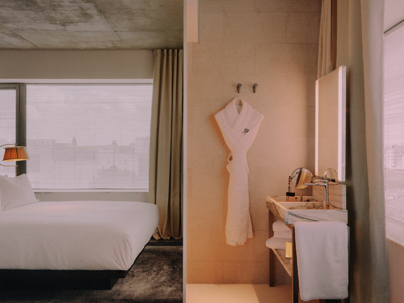A modern hotel room with a neatly made bed, large windows, and an adjacent bathroom featuring a hanging white bathrobe and a well-lit vanity.