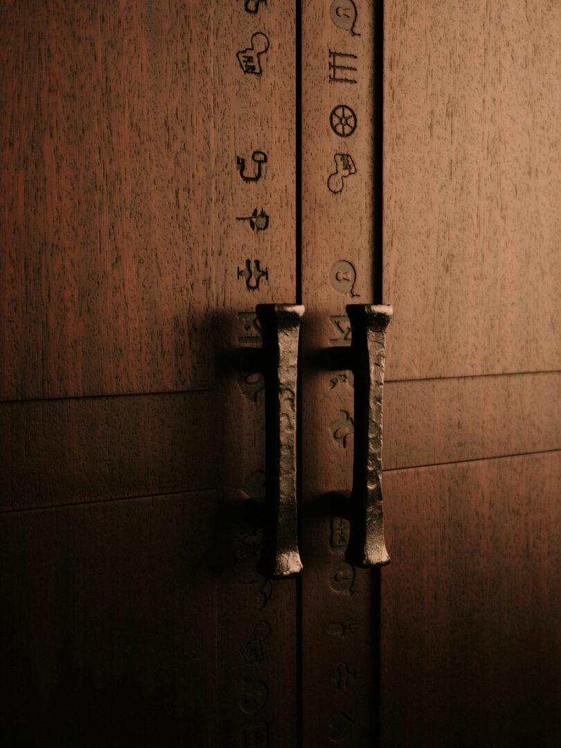 Close-up of two metal handles on a wooden cabinet door, with engraved symbols and characters arranged vertically along the seam between the doors.