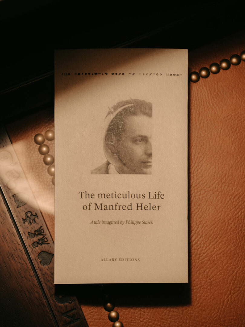 A book titled "The meticulous Life of Manfred Heler" with a sepia-toned cover featuring a portrait and text, placed on a leather surface next to a beaded item and ruler.