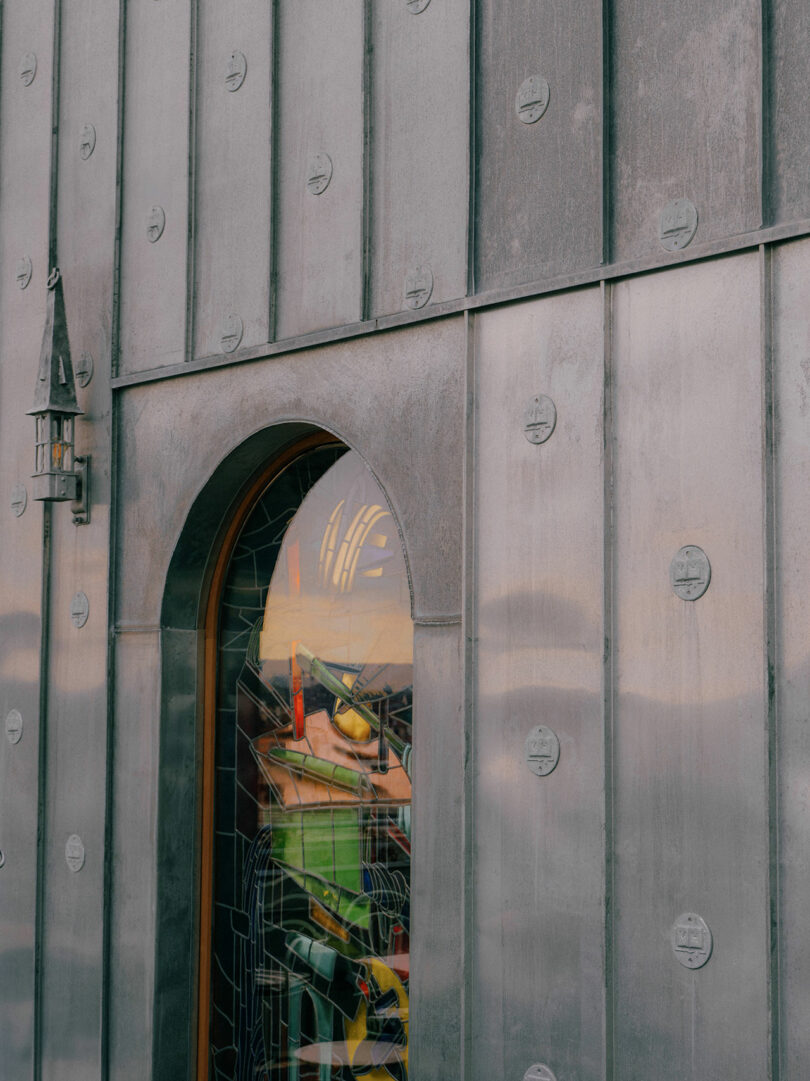 A metal-paneled building facade with a rounded arch window reflecting the sky and a lantern mounted on the wall beside the window.