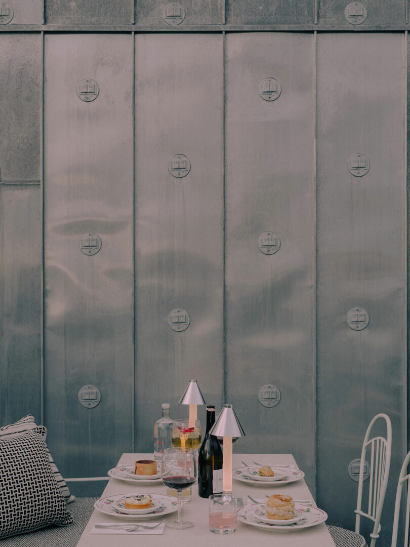 A set dining table with plates of pastries, drinks, and table lamps in front of a metallic wall; pillows and chairs are arranged around the table.