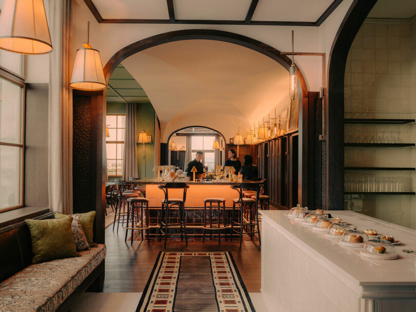 A warmly lit restaurant interior with a central bar, wooden chairs, arched doorways, and a display counter with pastries in glass domes. Two people stand behind the bar.