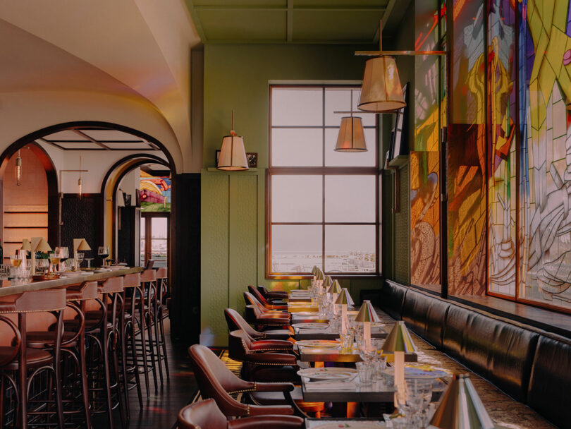A modern restaurant interior with neatly set tables, leather chairs, a bar area, large windows, and colorful stained glass wall art.
