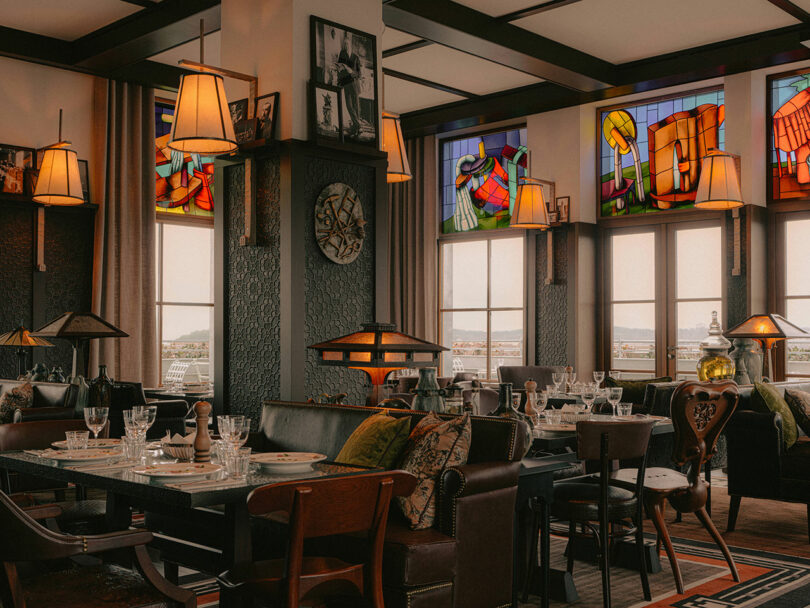 A stylish restaurant interior with set dining tables, cushioned chairs, stained glass windows, and warm, ambient lighting.