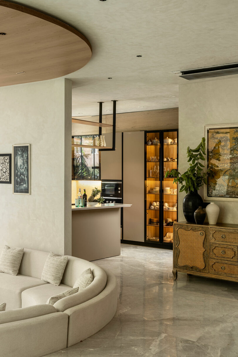 Modern living room with a curved white sofa, marble floor, and view into a kitchen with glass cabinets. Decorative plants and framed art accent the neutral-toned space.