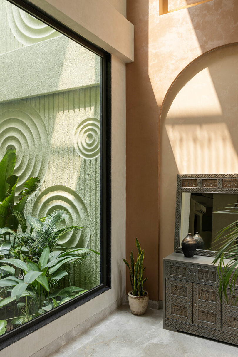 Sunlit indoor space with a large window, geometric wall art outside, potted plants, and an ornate cabinet with a mirror and decorative vases.