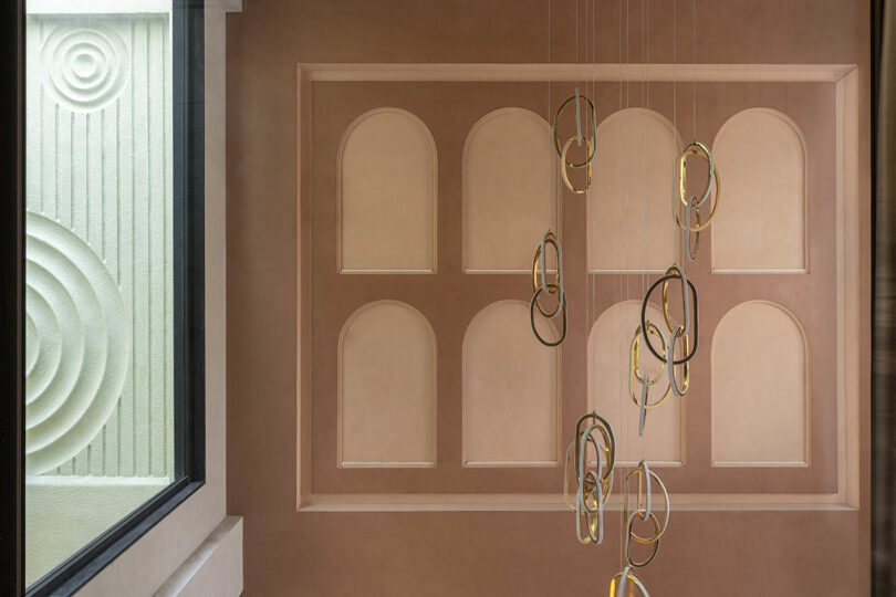 A modern interior with a wall featuring arched recesses, a sculptural gold ring chandelier, and a window showing a textured surface with concentric circular patterns.