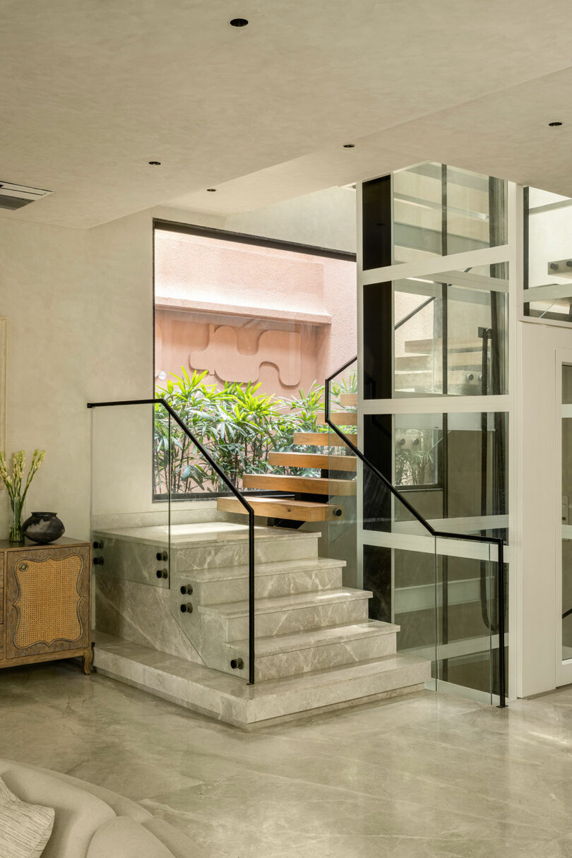 A modern indoor staircase with glass railings and marble steps, adjacent to a large window and a wooden cabinet; greenery is visible outside.