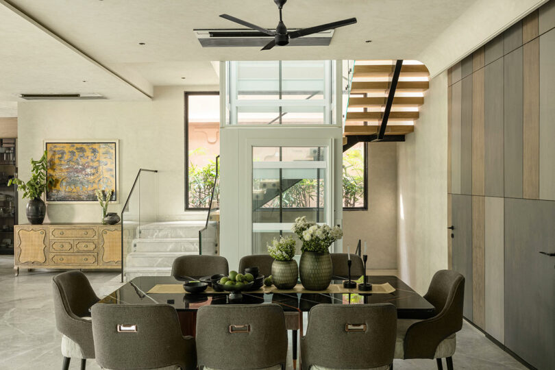 Modern dining area with glass table, six chairs, decorative vases, and staircase beside large windows; artwork and sideboard are visible in the background.