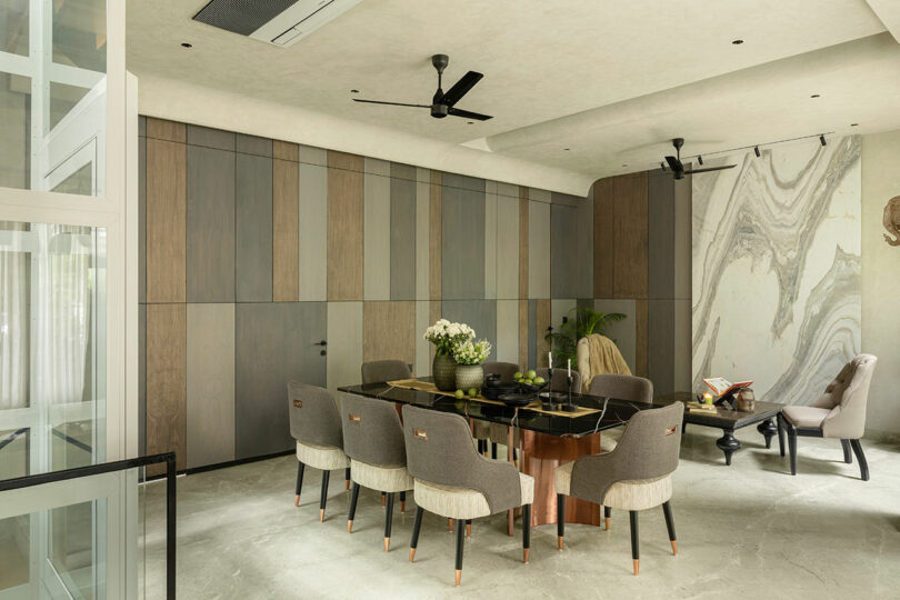 A modern dining room with a glass table, eight upholstered chairs, striped wood wall panels, and marble accent wall; a potted plant and decorative items complete the space.