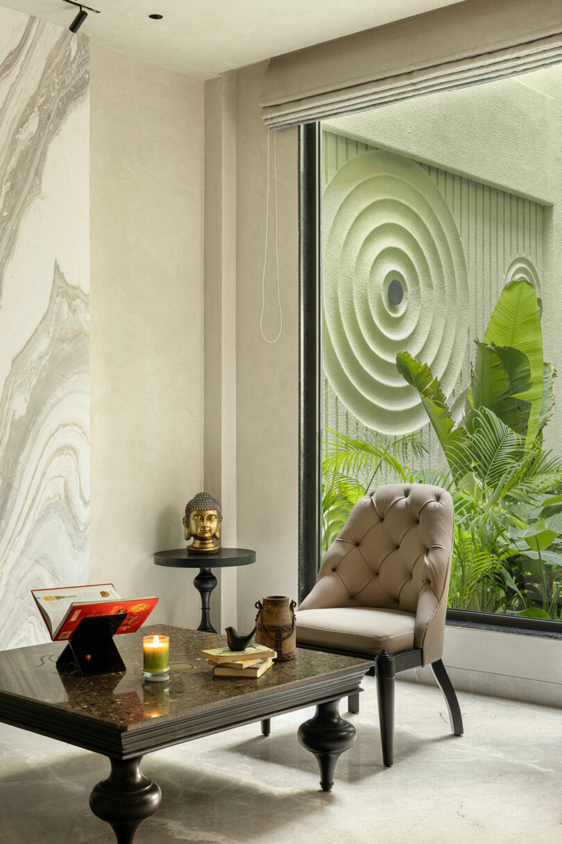 A modern room with a tufted chair, a dark coffee table with books and a candle, a small side table with a Buddha head, and a large window overlooking green plants and a textured wall.