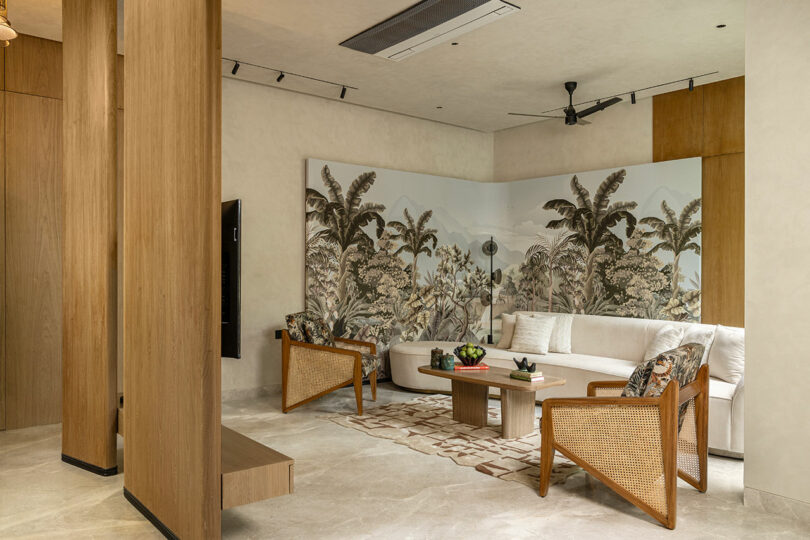 A modern living room with neutral tones, cane armchairs, a white sofa, a patterned rug, and a tropical-themed wall mural with palm trees.