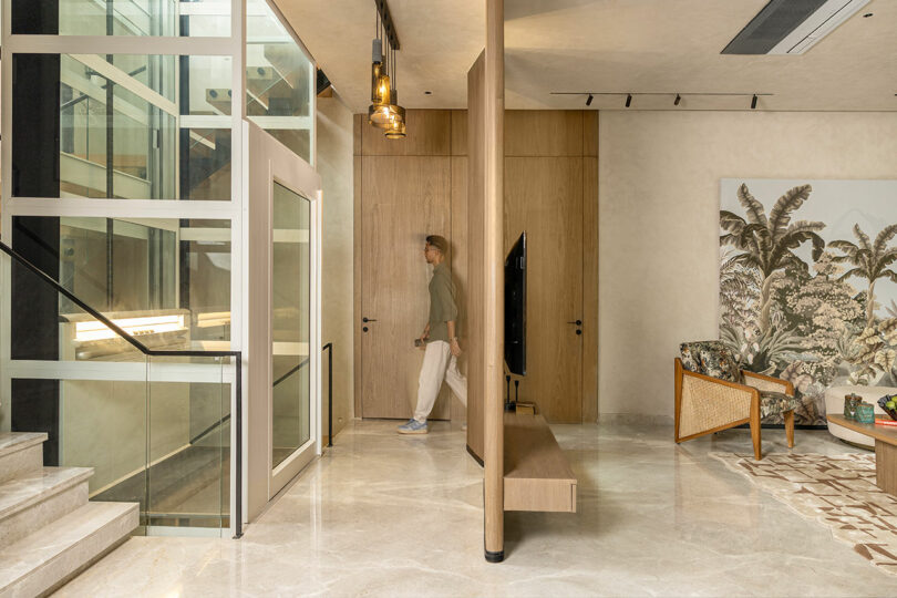 A man walks through a modern interior with marble floors, a glass elevator, wooden accents, and contemporary furniture.