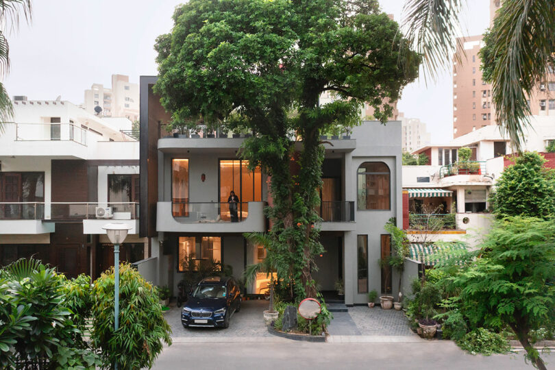 Modern two-story house with large tree integrated into the front entrance, a car parked in the driveway, and a person standing on the balcony.