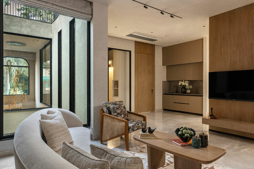 Modern living room with a curved sofa, armchairs, wooden coffee table, built-in TV, large windows, and minimalist decor in neutral tones.