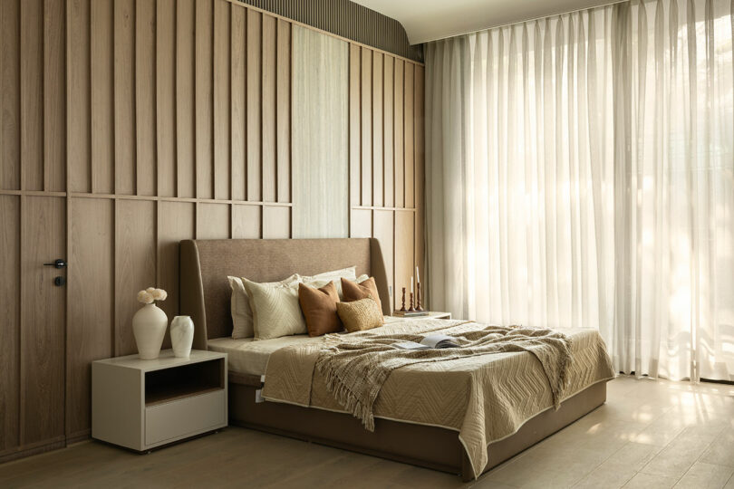 Modern bedroom with a large bed, neutral bedding, decorative pillows, a nightstand with vases, and floor-to-ceiling sheer curtains letting in natural light.