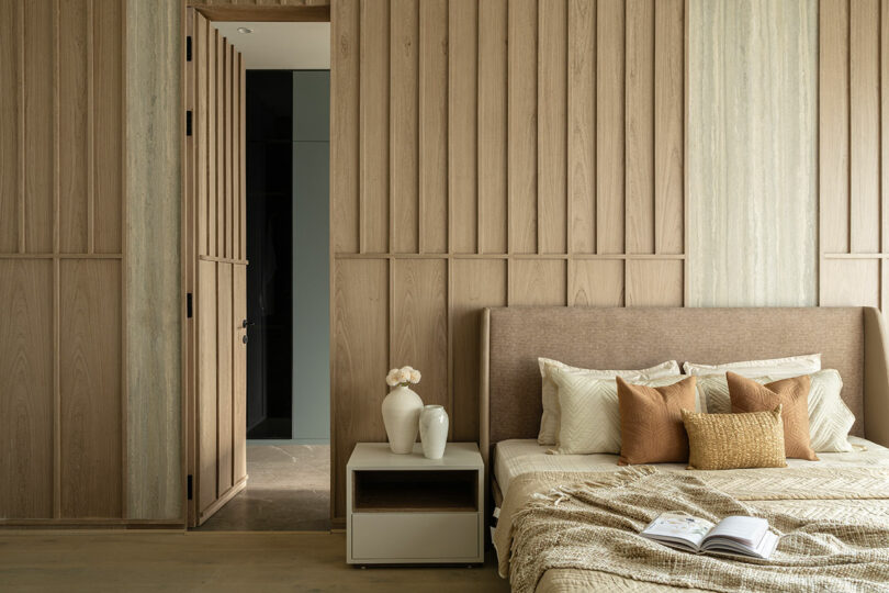 A modern bedroom with wood panel walls, a bed with neutral bedding and pillows, a nightstand with two white vases and a small flower arrangement, and an open door leading to a hallway.