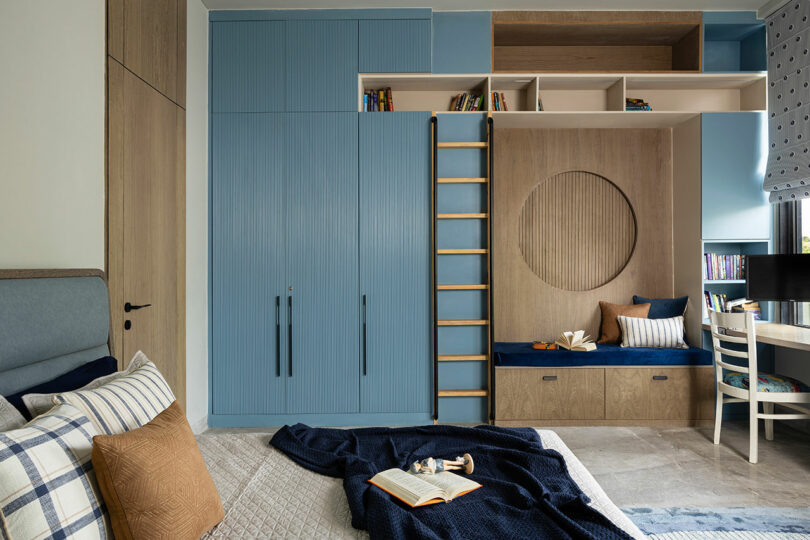 Modern bedroom with a bed, blue wardrobe, built-in shelving, a wooden ladder, window seat, desk, and neatly arranged pillows; open book on the bed.