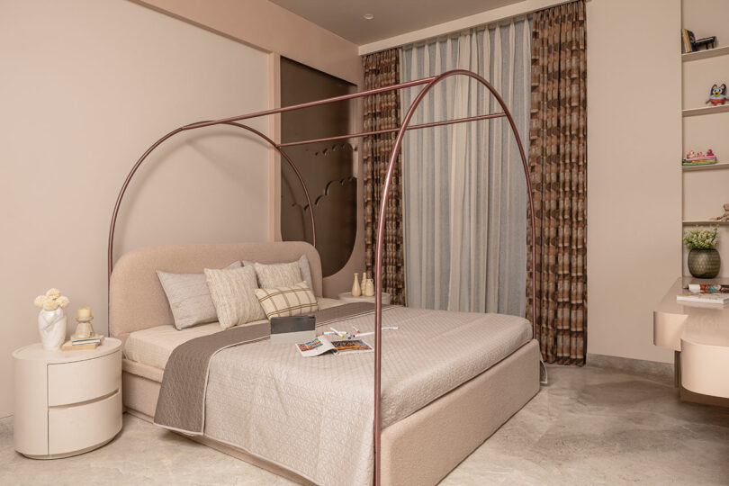 A modern bedroom with a beige canopy bed, neutral bedding, two round nightstands, books, and decorative items, against cream walls and floor-to-ceiling patterned curtains.