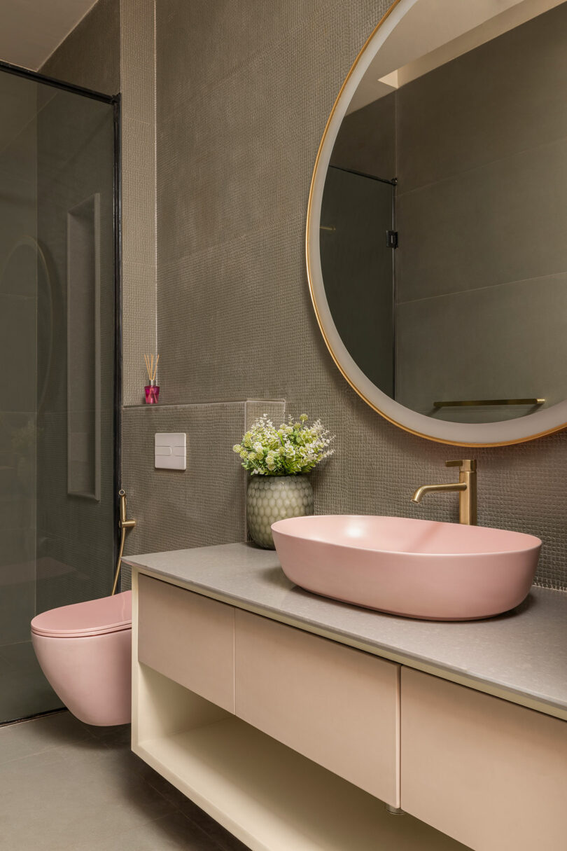 Modern bathroom with a round mirror, pink vessel sink, matching pink toilet, gold fixtures, open shelving, and a vase of white flowers on the countertop.