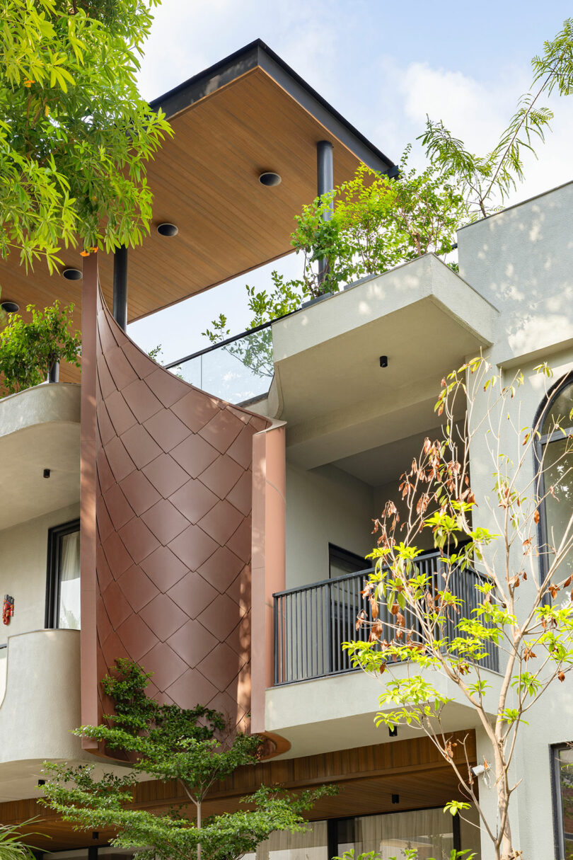 A modern building facade features a distinctive curved metal panel, balconies with black railings, rooftop greenery, and light gray walls surrounded by trees.