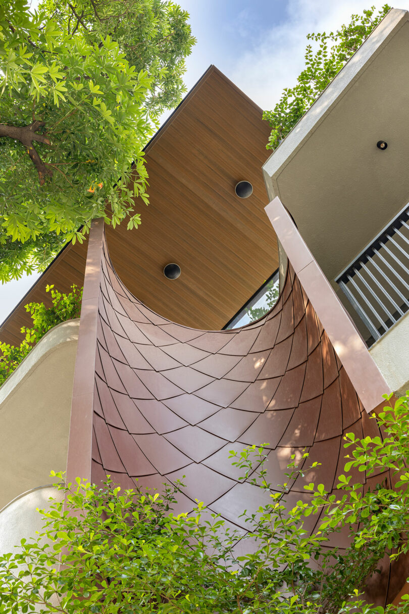 Low-angle view of a modern building facade featuring a curved, patterned metal panel, wooden ceiling detail, balcony railing, and greenery under a blue sky.
