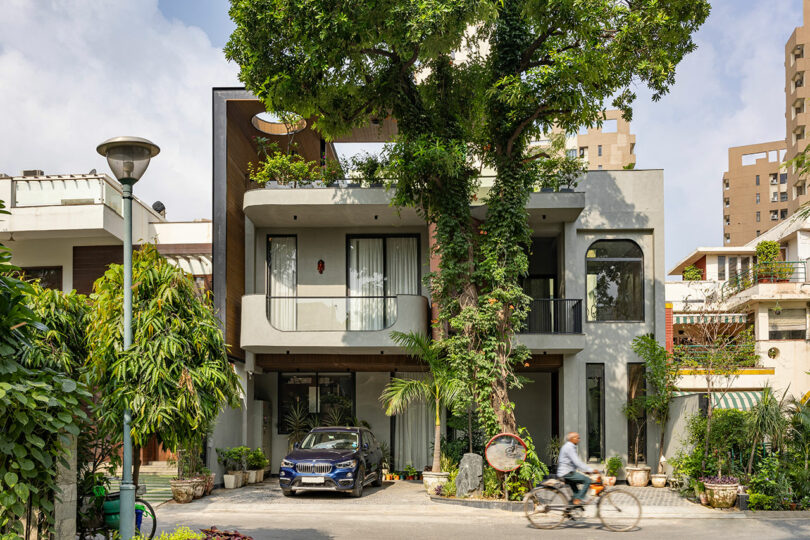 Modern two-story house with large tree integrated into the façade, surrounded by greenery, a parked car, and a person riding a bicycle in the foreground.