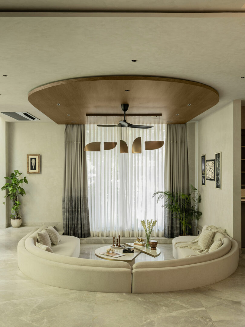 A modern living room with a curved beige sofa, round coffee table, houseplants, large windows with sheer curtains, and a ceiling fan under a wooden panel.