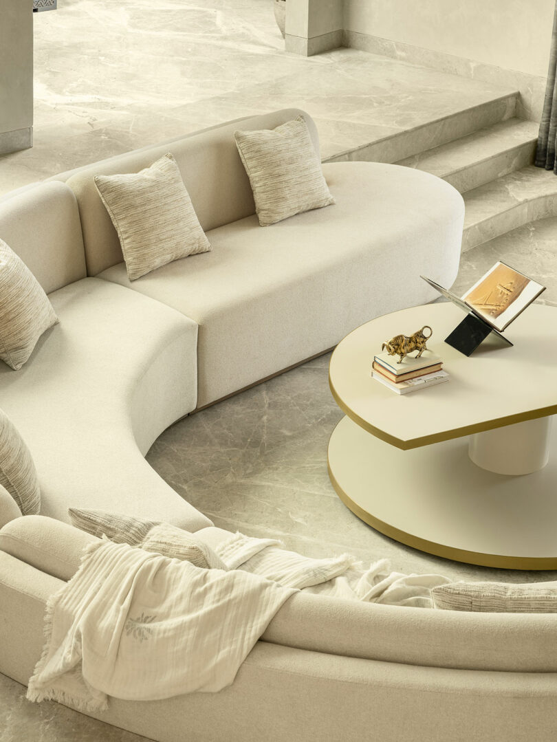 Curved beige sofa with neutral cushions and a white throw, surrounding a round coffee table with books, a small sculpture, and an open laptop on a light marble floor.