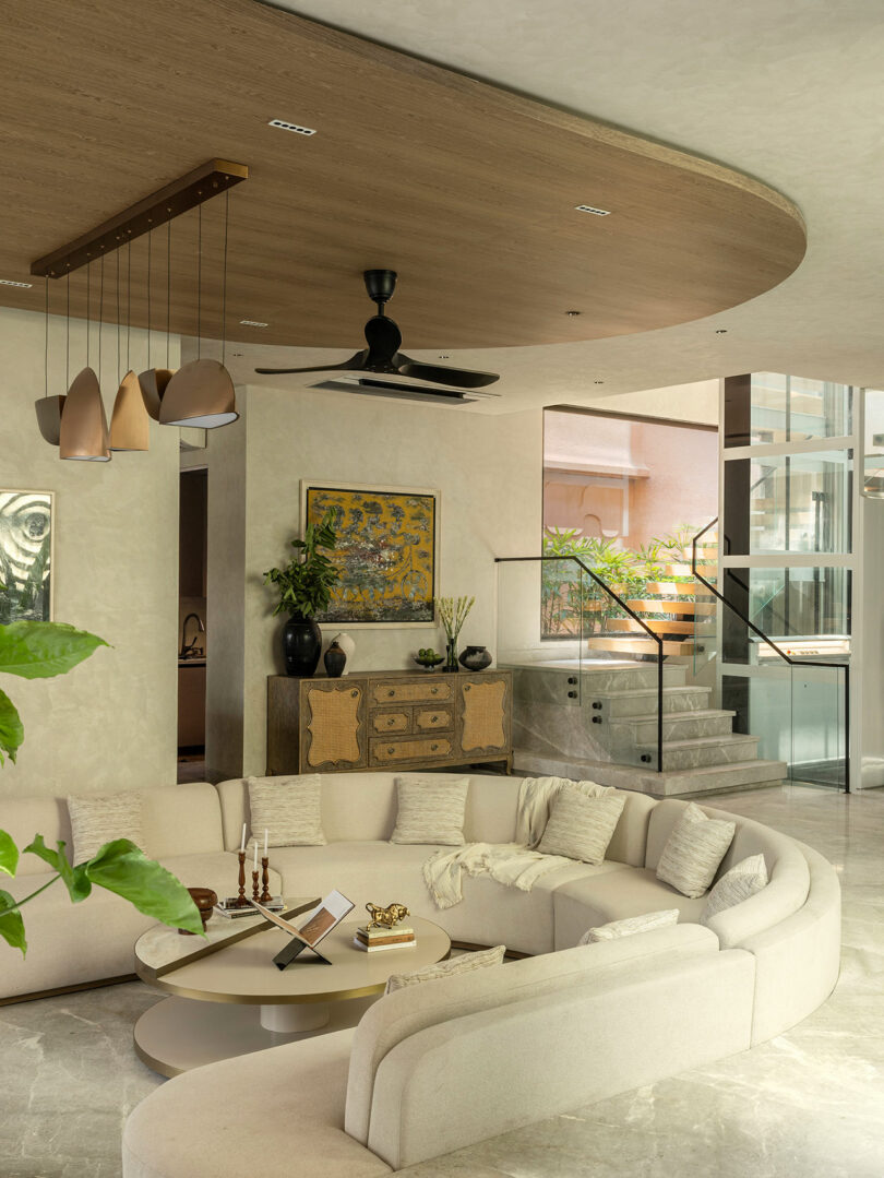 Modern living room with a curved beige sectional sofa, round coffee table, pendant lighting, ceiling fan, sideboard with decor, art on the wall, and stairs with a glass railing.