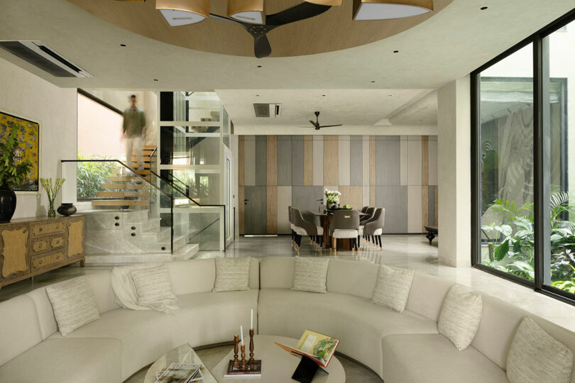 Modern living room with a curved beige sofa, glass coffee table, large windows, indoor plants, and a person walking down a staircase. Dining area is visible in the background.
