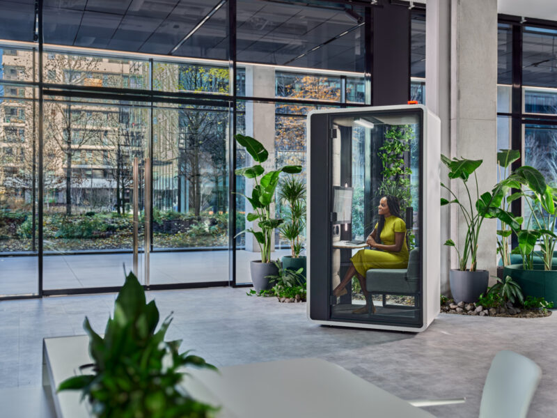 Woman in a lime green dress working inside a modern acoustic office pod surrounded by lush indoor plants.