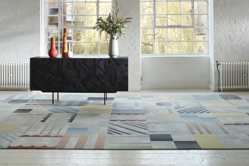 Colorful geometric carpet tiles in a bright room with a black textured sideboard and large windows.