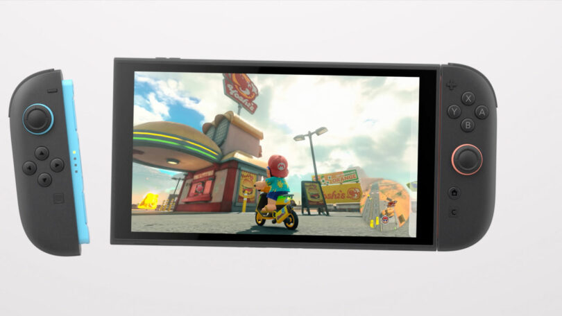 A Nintendo Switch 2 with blue and black Joy-Con controllers displays a Mario Kart game scene featuring a character on a motorcycle in front of a burger restaurant.