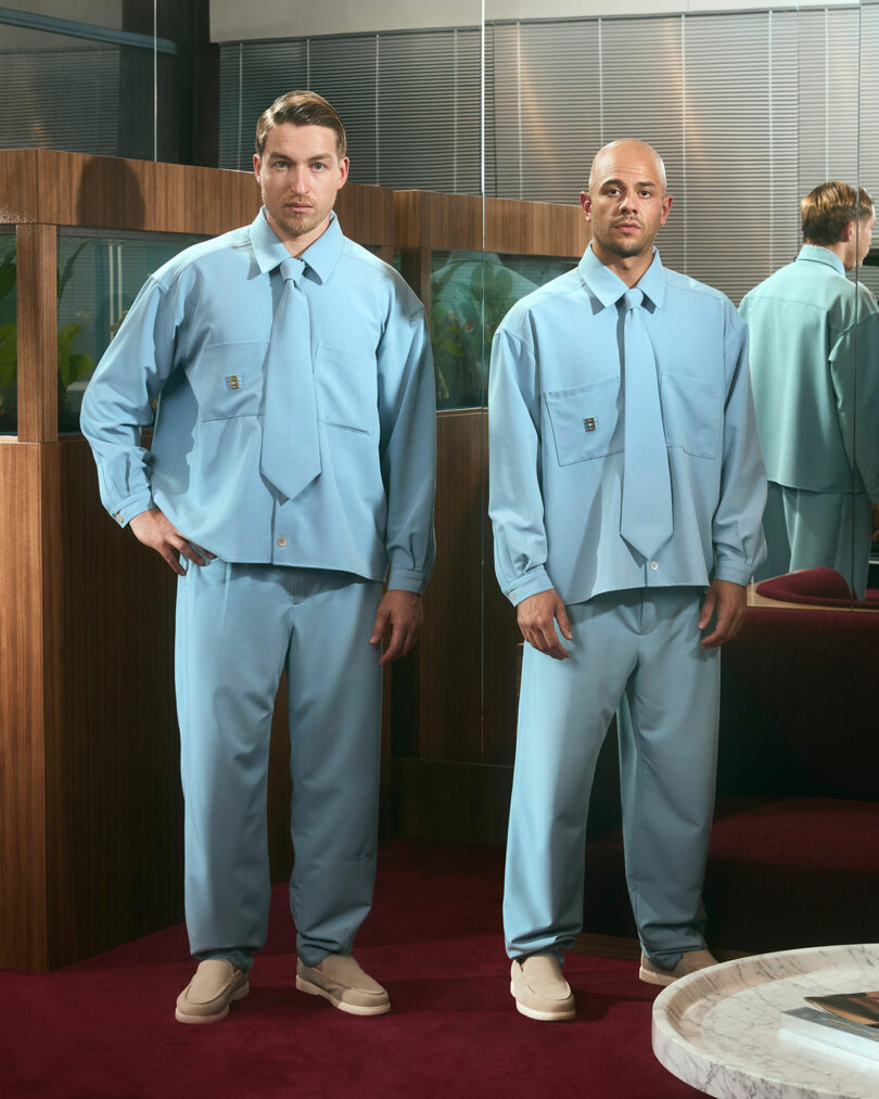Two men stand indoors wearing matching light blue suits with oversized shirts and ties, loose trousers, and beige shoes—a modern look reminiscent of the Futurespective Range Rover style. A third man is partially visible in the background reflection.