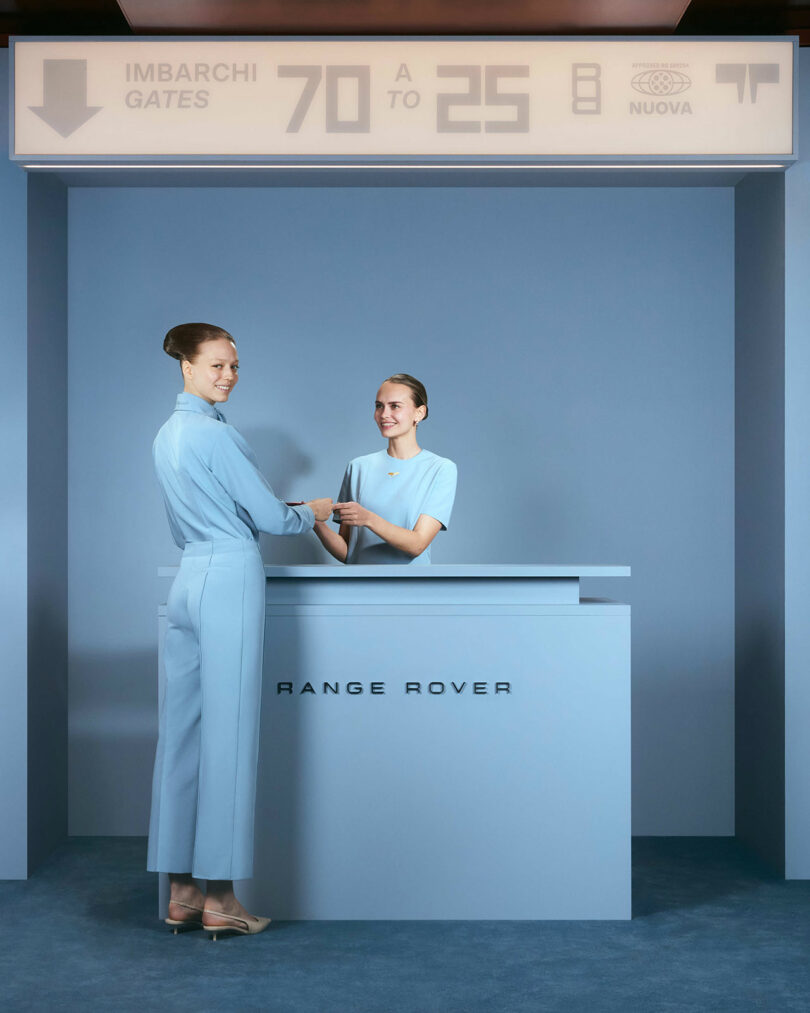 Two women in matching light blue outfits stand at a Futurespective Range Rover-branded counter in an airport setting, with directional gate signage above them.