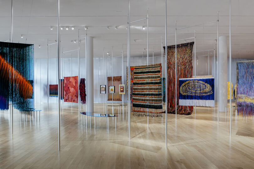 Large contemporary gallery with polished wood floors, displaying various colorful woven textile artworks hanging from thin vertical poles.