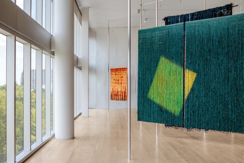 Three large hanging textile artworks displayed in a bright, modern gallery with floor-to-ceiling windows and light wood floors.