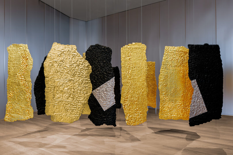 Several large, textured panels in gold, black, and silver hang suspended from the ceiling in a minimalist gallery space with wooden floors.